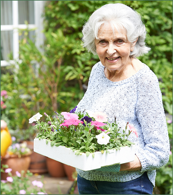 Photo of older woman standing outdoors holding a tray of flowers and smiling at the camera.